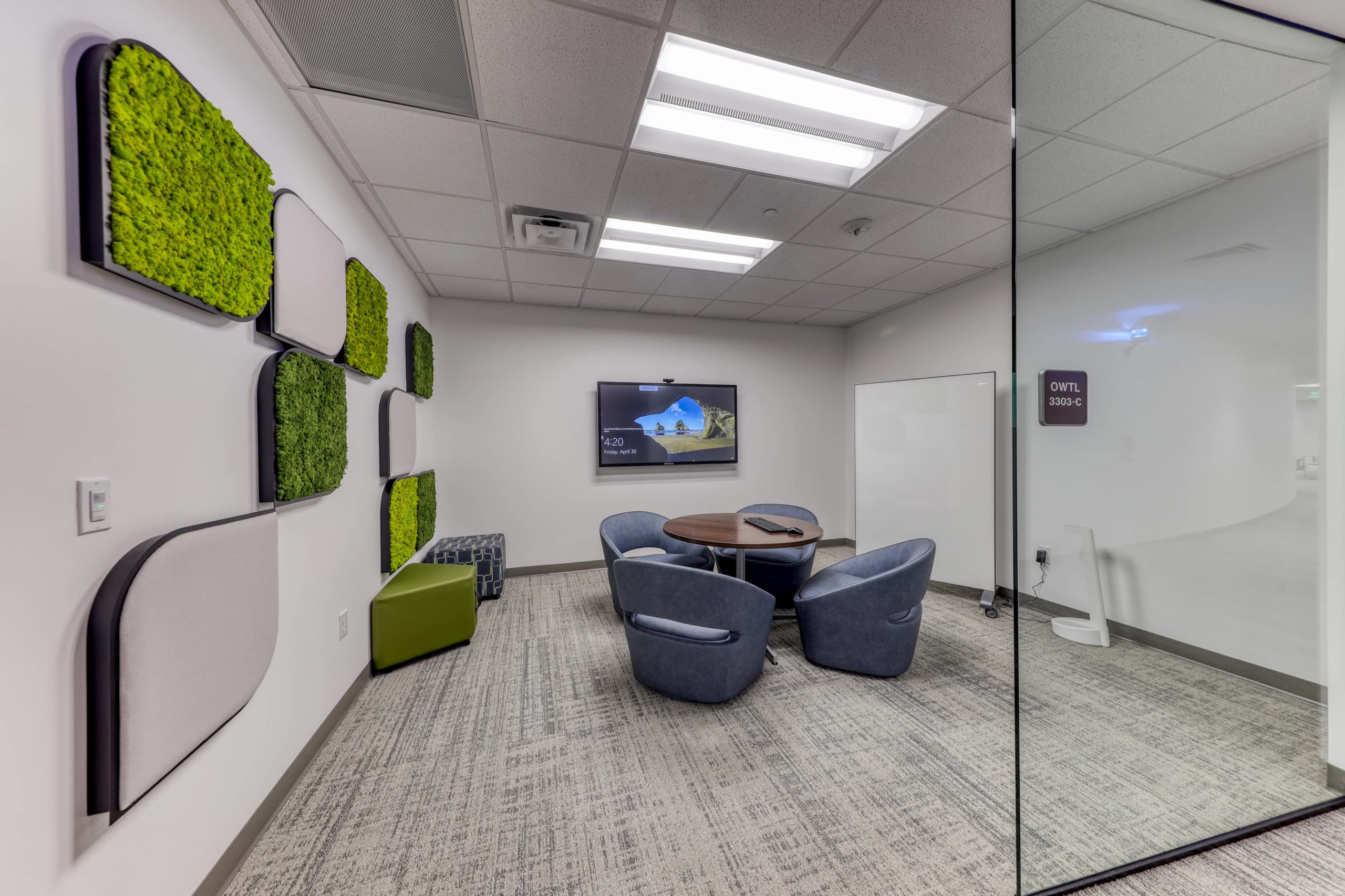 Glass-enclosed meeting room with round table, upholstered chairs, wall-mounted display, and acoustic wall tiles.