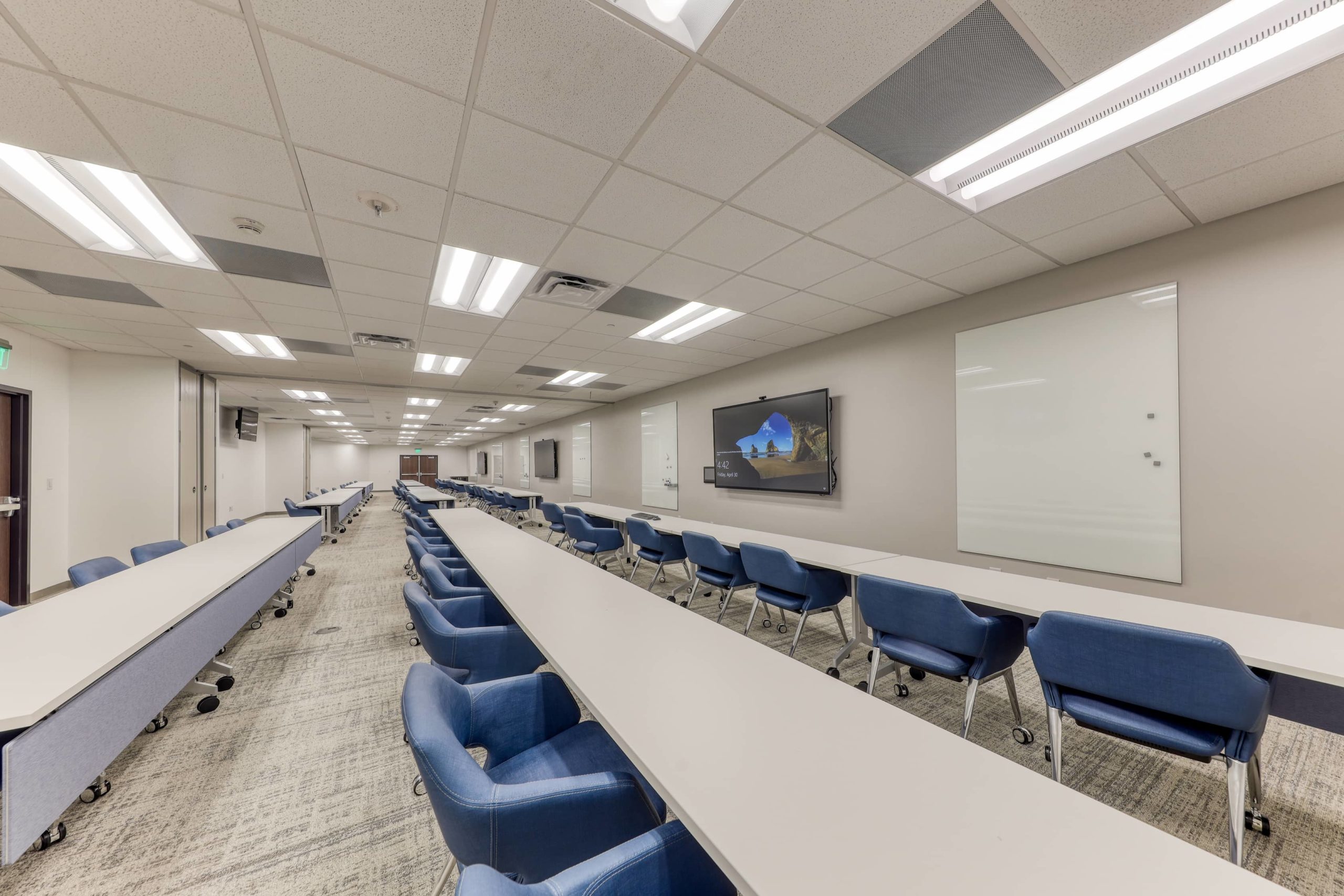 Large classroom with long tables, rolling chairs, wall-mounted displays, and writable surfaces for training sessions.