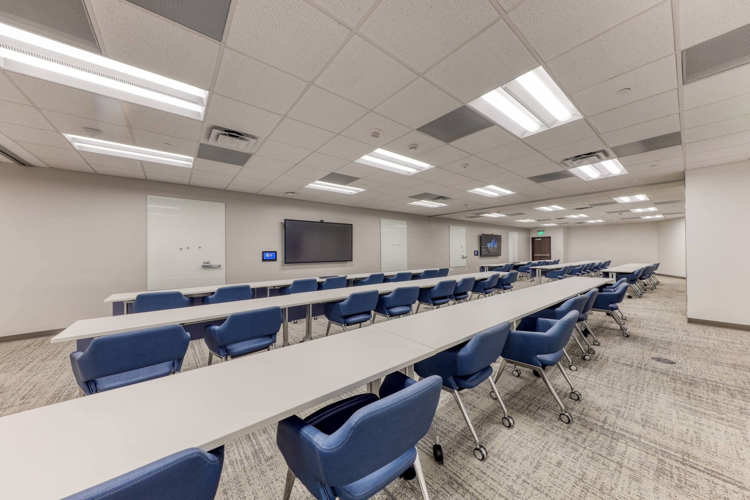 Extended training room with multiple rows of tables, mobile seating, presentation screens, and whiteboard walls.