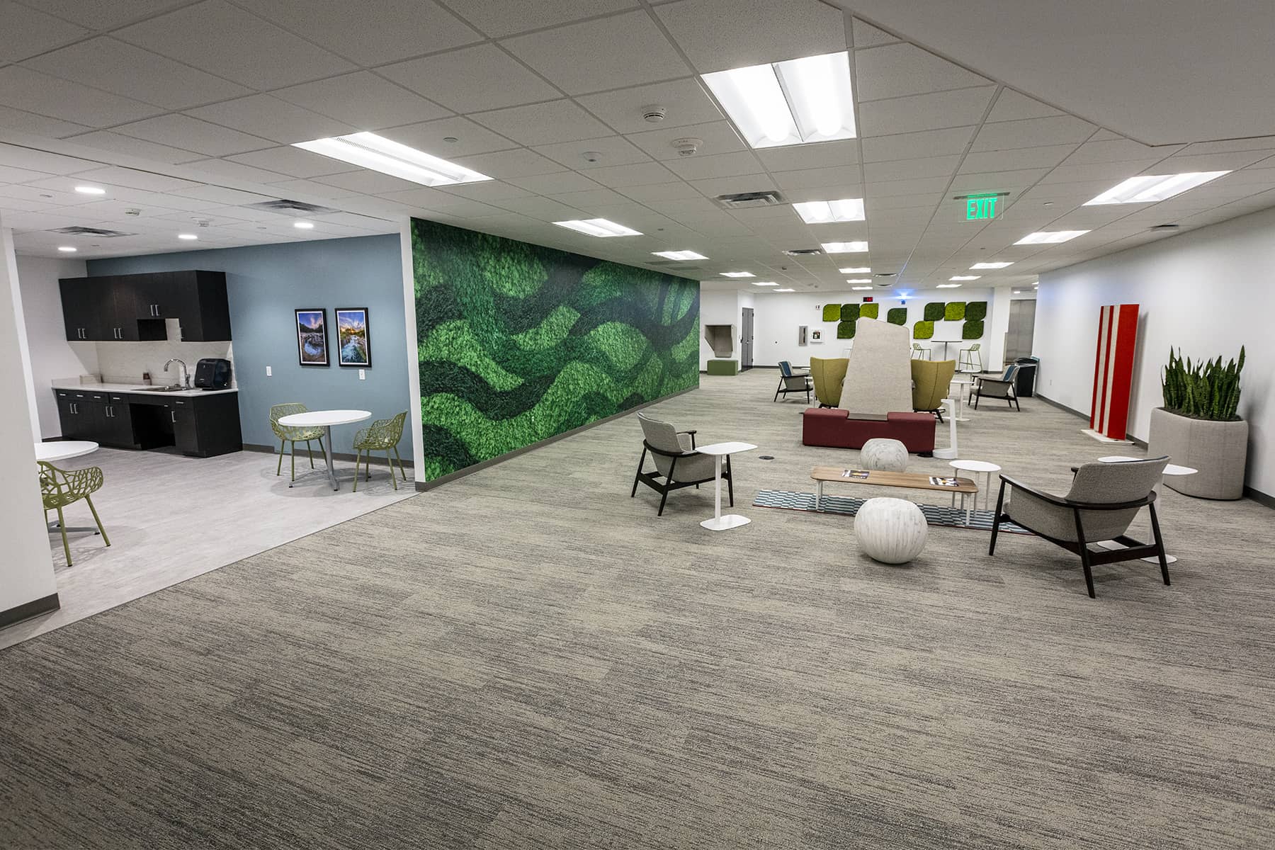 Wide view of a flexible common area with seating clusters, greenery wall accents, and an adjacent kitchenette.