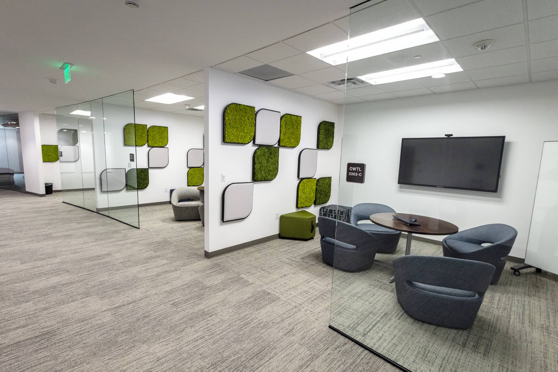 Glass-walled meeting room with circular table, upholstered chairs, wall-mounted display, and decorative acoustic panels.